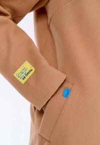 Bežový rukáv kabátu se žlutou nášivkou s nápisem "žádné limity" a modrou etiketu se zaklopenou řasou poblíž boční kapsy.