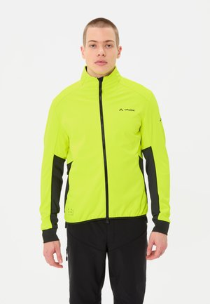 Junger Mann trägt eine leuchtend gelb-schwarze Radjacke mit Reißverschluss, steht frontal mit neutralem Gesichtsausdruck vor weißem Hintergrund.