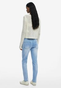 Pull en crochet crème avec un ourlet festonné, associé à un jean bleu clair avec poches arrière et coupe slim. Les chaussures sont de couleur gris clair.