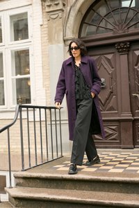 Lila strukturierte Jacke, schwarzes Rüschenshirt, weitgeschnittene schwarze Hose und schwarze Schuhe. Schauplatz: Steinstufen neben einer verzierten braunen Tür.