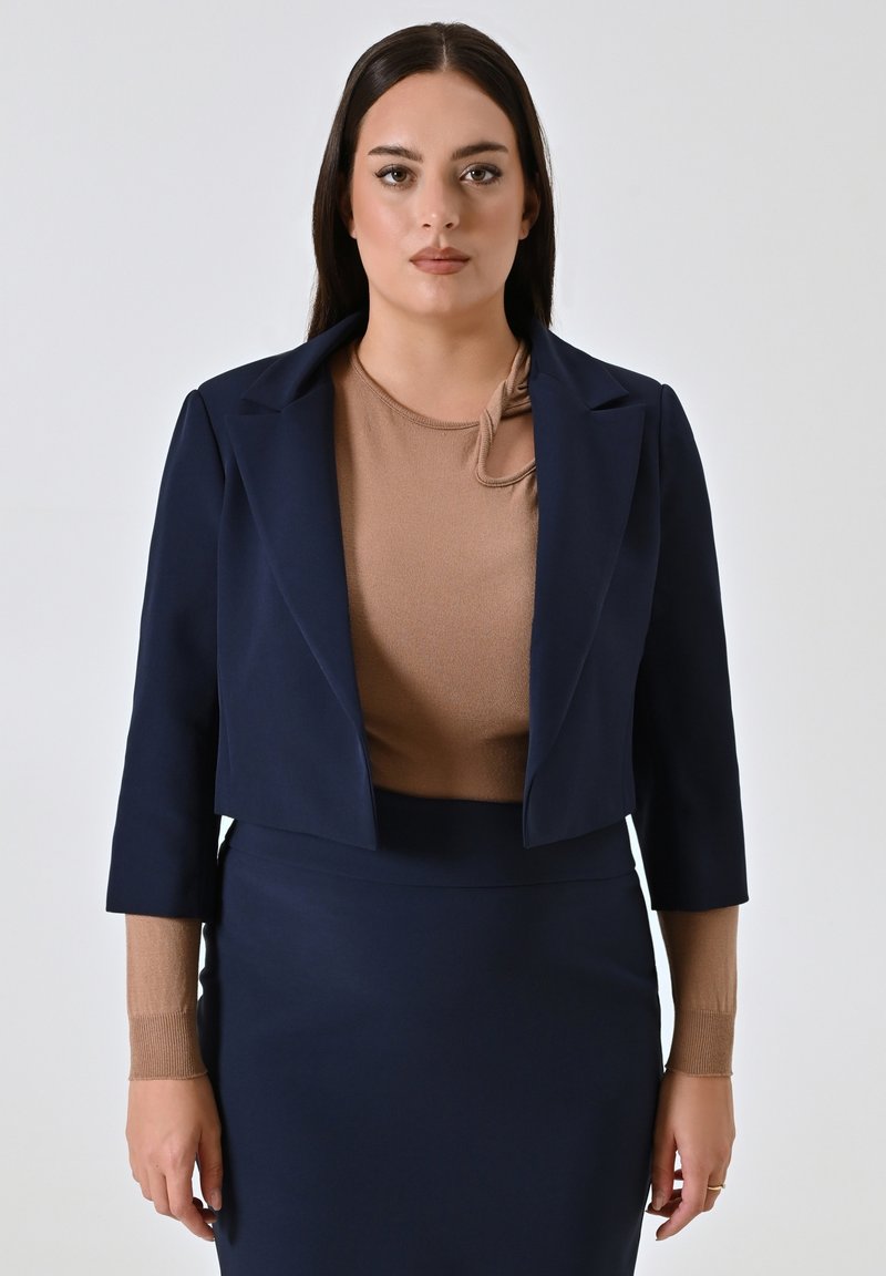 Marineblauw cropped blazer met gestructureerde schouders, gedragen over een beige top met knoopdetail. Soepele stof met een getailleerde pasvorm.