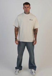 Kermanvärinen t-paita, jossa lyhyet hihat ja musta logo, yhdistettynä löysiin sinisiin farkkuihin ja valkoisiin lenkkareihin. Malli seisoo yksiväristä taustaa vasten.