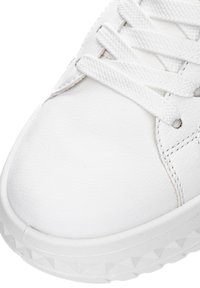 Detailní záběr na špičku bílé tenisky s texturovanou kůží, bílými tkaničkami a vzorovanou gumovou podrážkou.