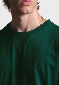 Zelené bavlněné tričko s krátkým rukávem, kulatým výstřihem a vyšitým logem slona na hrudi. Měkká, hladká textura.
