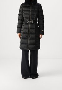 Abrigo largo negro acolchado tipo puffer con cuello de piel, cierre de cremallera, cinturón elástico con hebilla plateada, combinado con pantalones negros de pierna ancha y zapatos negros.