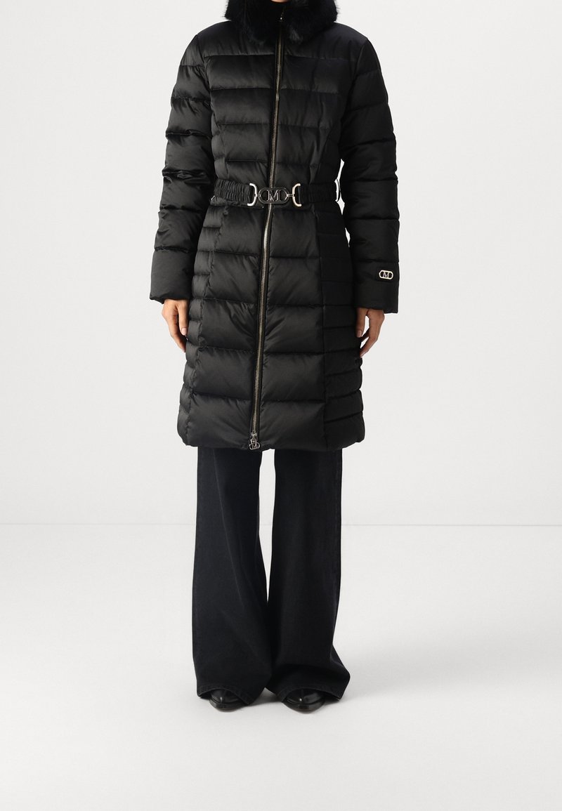 Abrigo largo negro acolchado tipo puffer con cuello de piel, cierre de cremallera, cinturón elástico con hebilla plateada, combinado con pantalones negros de pierna ancha y zapatos negros.