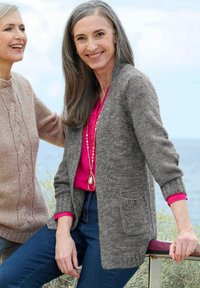 Lächelnde reife Frauen im Freien am Wasser, eine in einem grauen Cardigan und rosa Bluse, die andere in einem beigen Strickpullover, die sich unterhalten.