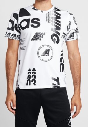 Hombre con camiseta blanca de Adidas con logos gráficos en negro audaz y texto, combinada con pantalones negros de Adidas con logo cerca del bolsillo.