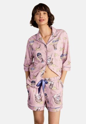 Femme portant un ensemble de pyjama rose avec des illustrations de montgolfière et de voilier, chemise boutonnée et short à cordon.