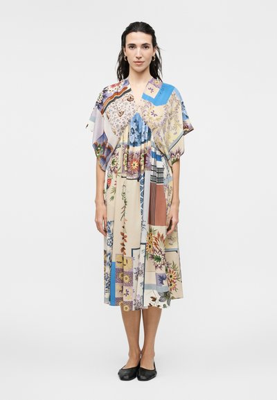 Femme debout portant une robe midi ample à motifs avec manches courtes et chaussures plates noires sur un fond clair uni.
