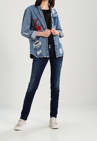 Giacca di jeans con ricami floreali rossi, dettagli strappati, abbinata a una maglietta nera e jeans blu scuro, completata con sneakers bianche.