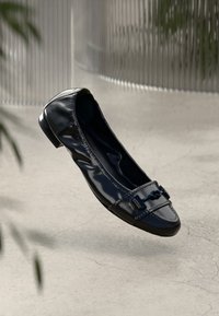 Chaussure plate en cuir verni noir avec un élément décoratif en métal sur le devant, présentée sur un fond texturé gris clair.