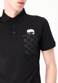 Svart polo skjorte med treknaps placket, som har en kontrastlomme med et geometrisk mønster og en hvit grafisk logo over lommen.