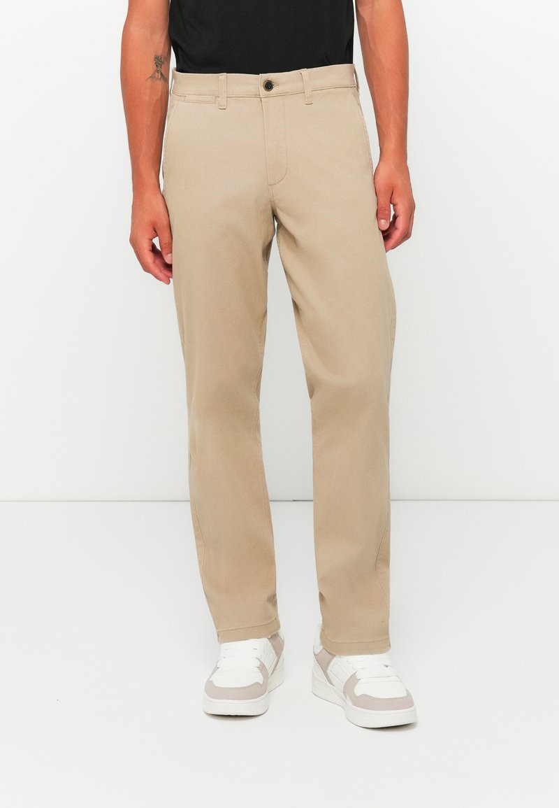 Chinos beige à coupe droite, fermeture à bouton et poches avant, associés à des baskets blanches. Le tissu semble lisse et résistant.