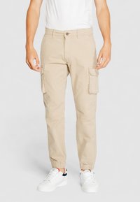 Beige Cargo-Hosen mit schmal zulaufenden Knöcheln, zwei Seitentaschen und elastischen Bündchen, aus leichtem Stoff, getragen mit weißen Sneakers.