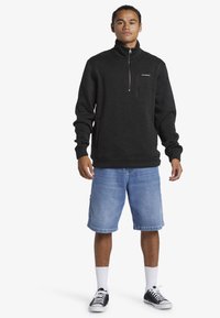 Čierny fleecový pulóver s štvrtinovým zipsom, rebrovanými manžetami a vreckom na hrudi. Skombinovaný s svetlomodrými džínsovými šortkami a čiernymi teniskami.