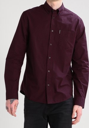 Homme portant une chemise à manches longues bordeaux et noire à carreaux avec une poche poitrine, associée à un pantalon noir.