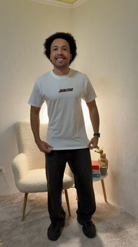 Man die lacht en een wit "Santa Cruz" T-shirt rechtzet, staand in een gezellige kamer met een fauteuil, een bijzettafel, boeken, een vaas en een kopje.