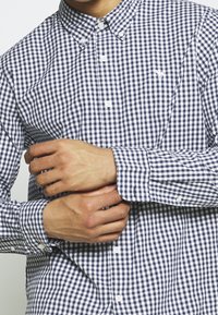 Niebieska i biała koszula w kratę typu gingham z kołnierzykiem zapinanym na guziki, długimi rękawami i białymi guzikami. Posiada małe logo na klatce piersiowej.