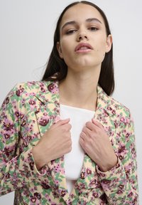 Blomstret blazer i creme, med lyserøde og grønne mønstre. Med jakkeslag, let tætsiddende design, lange ærmer og en tekstureret overflade.