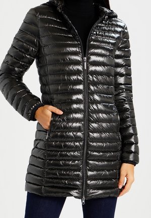 Femme portant une doudoune matelassée noire brillante avec une fermeture éclair devant et les mains dans les poches, associée à un pantalon foncé.