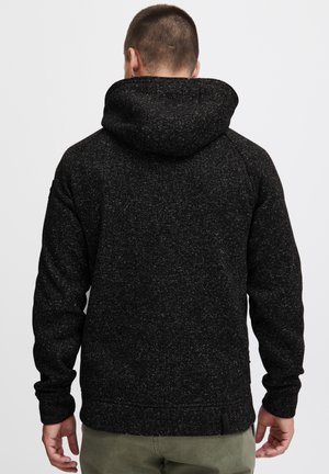 Sudadera con manchas negras y acabado texturizado, que presenta una capucha forrada, mangas largas y un corte relajado. Un detalle adicional incluye un pequeño cierre lateral.