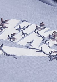 Ανοιχτού μπλε φούτερ με λευκό λογότυπο Nike που έχει ενσωματωμένα σκούρα μπλε και μωβ λουλουδάτα κεντήματα στο σχέδιο.