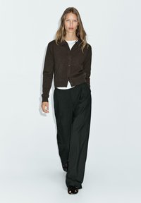 Cardigan marrone con bottoni, camicia bianca sotto e pantaloni larghi a righe verticali neri. Il modello indossa sandali neri con cinturini.