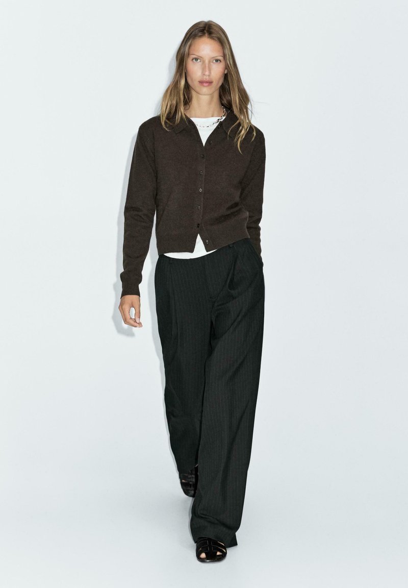 Brązowy kardigan z guzikami, biała koszula pod spodem oraz czarne spodnie w szerokie paski. Modelka nosi czarne sandały na paski.