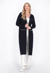 Marineblauer langer Cardigan mit weißem Saum, offener Front, zwei aufgesetzten Taschen und einem leicht strukturierten Stoff, getragen über hellen Hosen und Sneakern.