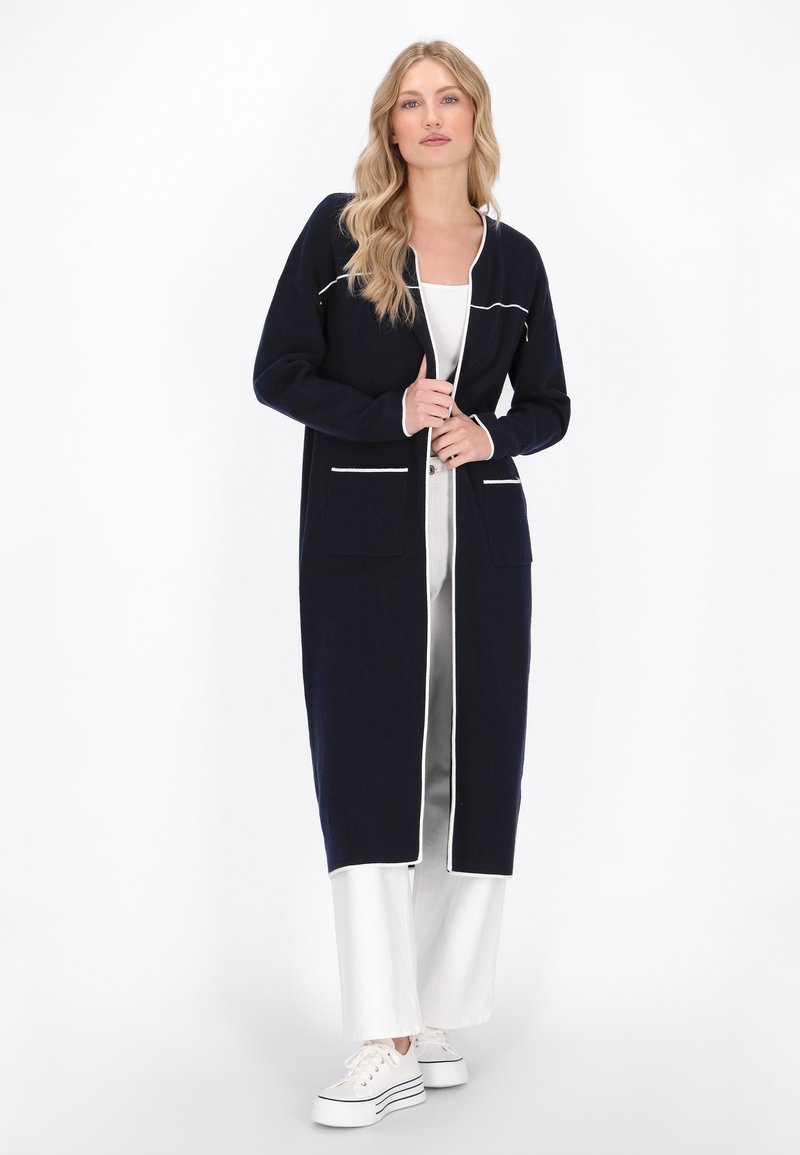 Cardigan lung de culoare albastru marin cu margine albă, deschis în față, cu două buzunare aplicate și un material ușor texturat, purtat peste pantaloni deschiși la culoare și adidași.