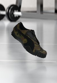 Camouflage-patterned sports shoe with black textured sole floating above a gym floor with a blurred dumbbell in the background.