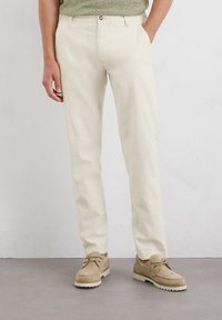 Cream-colored trousers made of lightweight fabric, featuring a straight leg design, side pockets, and a button closure, paired with beige shoes.