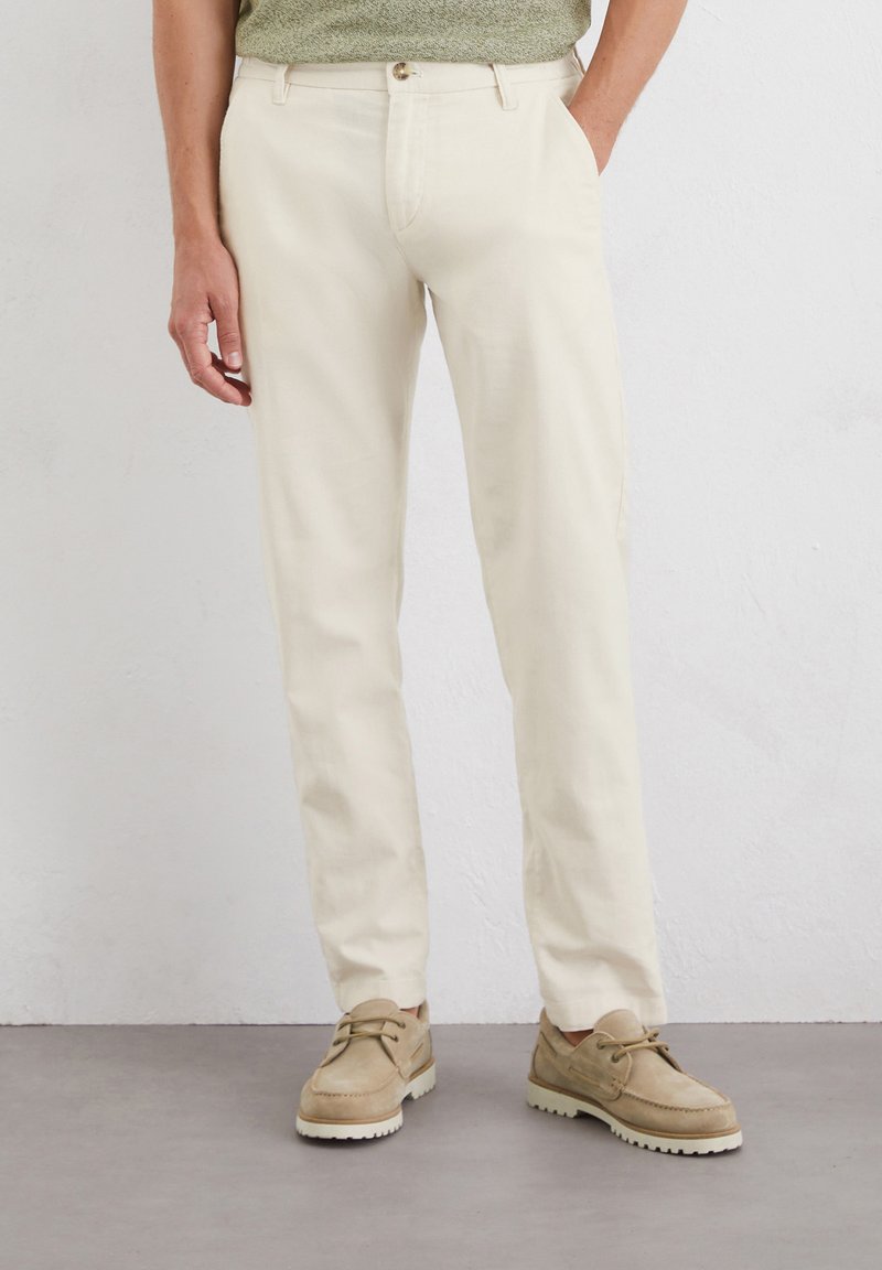 Cream-colored trousers made of lightweight fabric, featuring a straight leg design, side pockets, and a button closure, paired with beige shoes.