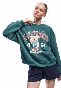Teal sweatshirt med oversized passform, med en björn grafik och texten "CALIFORNIA 1964". Kombinerad med mörkblå shorts. Slät tygtextur.