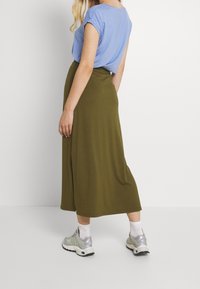 Donne in piedi di lato che indossa una maglietta azzurro chiaro, una gonna midi verde oliva, calzini bianchi e scarpe da ginnastica grigie su uno sfondo neutro.