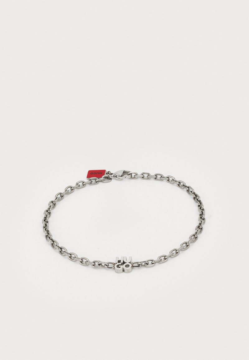 Pulsera de cadena de plata con eslabones entrelazados y un detalle central del logo "HUGO". Incluye una pequeña etiqueta de marca roja y un cierre de broche.