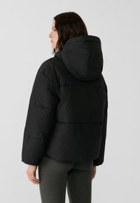 Doudoune noire avec capuche, présentant un design oversized, une texture matelassée et des poignets élastiqués. La vue arrière montre la longueur raccourcie.