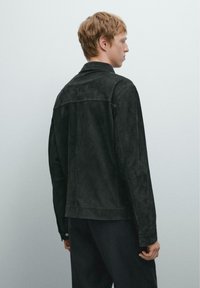 Schwarze Wildlederjacke mit langen Ärmeln, geradem Schnitt und minimalen Details. Von hinten gezeigt, mit einer sauberen, strukturierten Oberfläche.