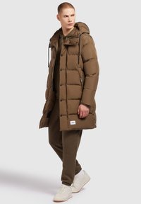 Bruine pufferjas met capuchon, voorzien van een doorgestikt ontwerp en ritszakken. Draagt over olijfgroene joggers en witte sneakers.