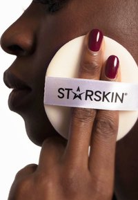 Tampon de nettoyage rond et blanc tenu entre des doigts vernis de rouge profond. Il présente une banderole blanche avec "STARSKIN" imprimé en noir.