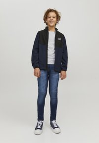 Giacca di felpa blu navy con pannelli neri, t-shirt bianca, jeans blu e sneakers blu navy. Logo ricamato sul lato superiore sinistro della giacca.