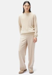 Beige stickad tröja med rund hals, ihop med ljusbeige lösa byxor. Modellen bär mjuka, platta bruna skor. Enkelt, strukturerat mönster.