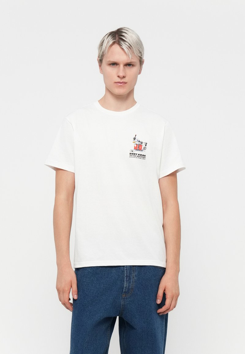 White cotton t-shirt with a round neck and short sleeves. Features a colorful graphic design on the left chest. Worn with blue denim jeans.
