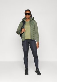 Gilet verde imbottito sopra una maglietta a maniche lunghe verde, abbinato a leggins neri con tasche decorative e scarpe da trekking nere.