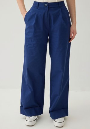 Person trägt hoch taillierte, weit geschnittene blaue Hosen mit aufgekrempelten Säumen und weißen Sneakers, Hände teilweise in den Taschen, steht vor einfarbigem Hintergrund.