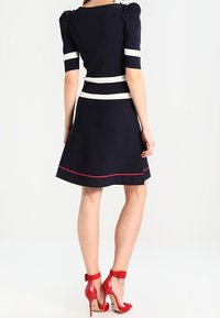 Femme portant une robe marine avec des rayures blanches sur les manches et la taille, bordure rouge près de l'ourlet, associée à des chaussures à talons hauts rouges avec bride à la cheville, vue de dos.