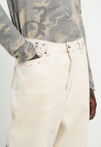 Jeans din denim bej cu croi relaxat, prezentând ușoare deteriorări și stropi de vopsea multicolori aproape de buzunare și cusături.