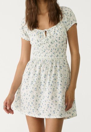 Kvinde iført kort hvid kjole med blå blomstermønster, smocktalje, ballonærmer og bindedetalje foran.