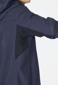 Marinblå jacka med en texturerad huva och ventilationpaneler under armarna med perforeringar, tillverkad av ett lätt, vattenavvisande material.